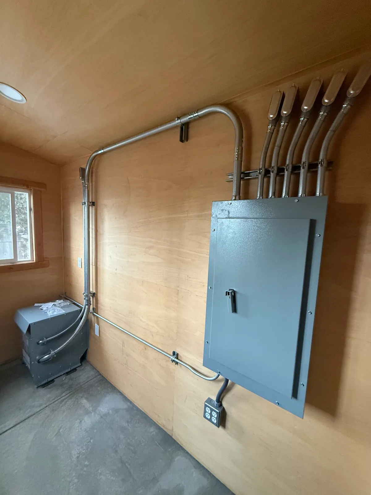 Panelboard with conduit runs and a transformer in a wood-paneled electrical room at the LA County Arboretum greenhouses