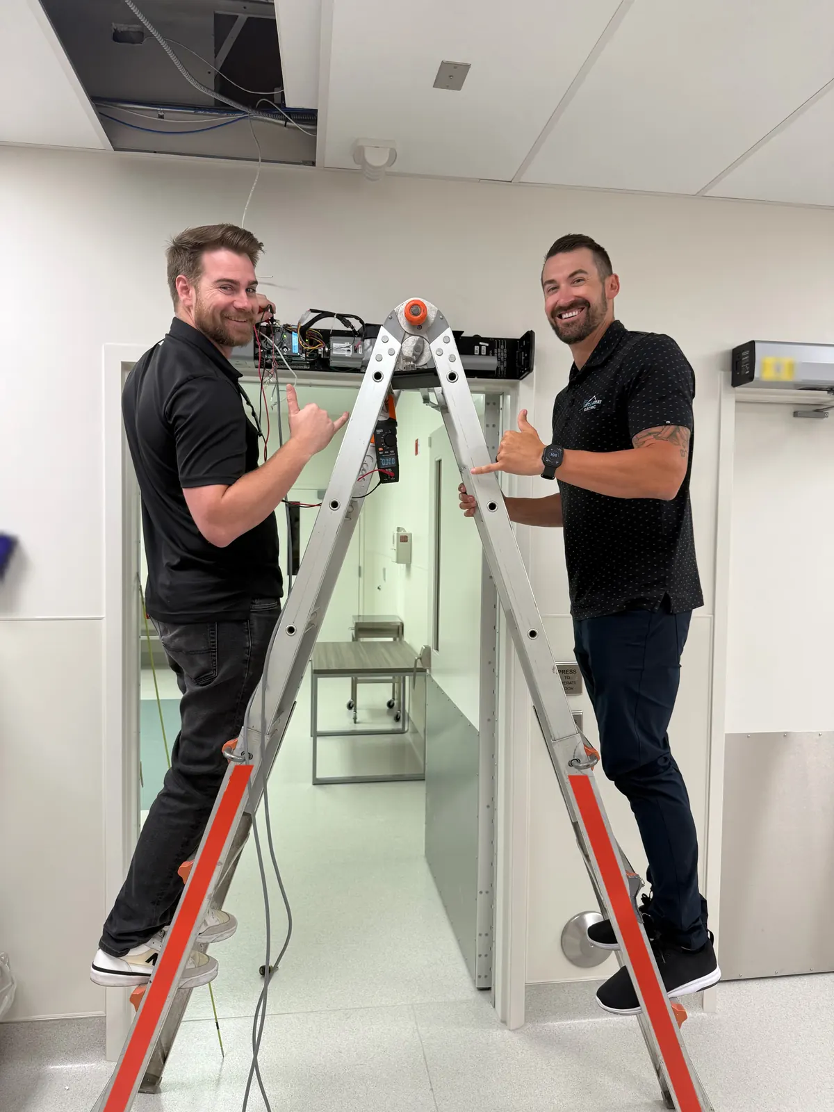 Two Left Coast Electric team members on a ladder at a doorway header with an open access-control panel at Ortho AZ in Glendale, AZ